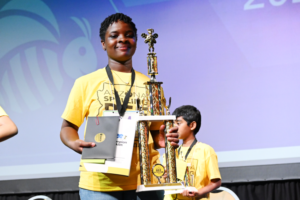 A participant in a yellow shirt holds a large trophy on stage at a spelling bee event, with another participant standing in the background.