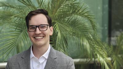 Man with glasses smiling, wearing a gray suit and white shirt, standing in front of green palm plants.