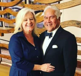 Two smiling individuals standing together, both wearing formal attire. One is in a dark suit, while the other is in a light blazer. They are indoors with a decorative screen in the background.
