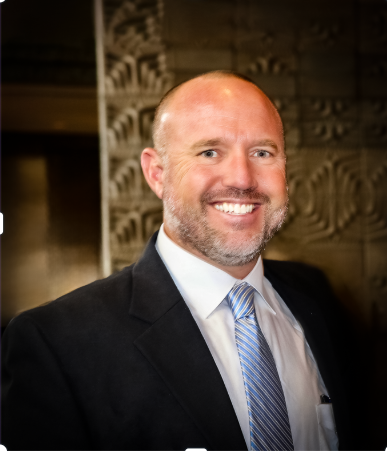 A man in a suit and tie smiles at the camera against a textured background.