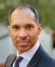 A person wearing a suit and tie smiles at the camera in an outdoor setting.