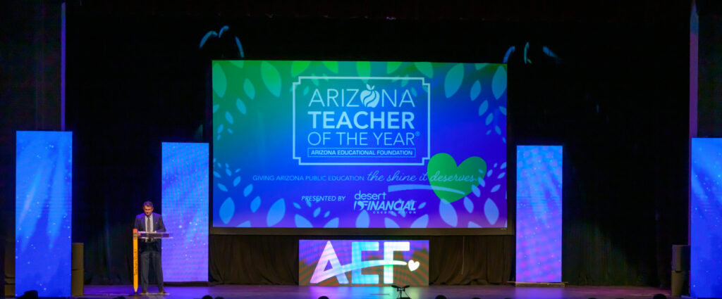 A person stands at a podium on stage next to a large screen displaying 