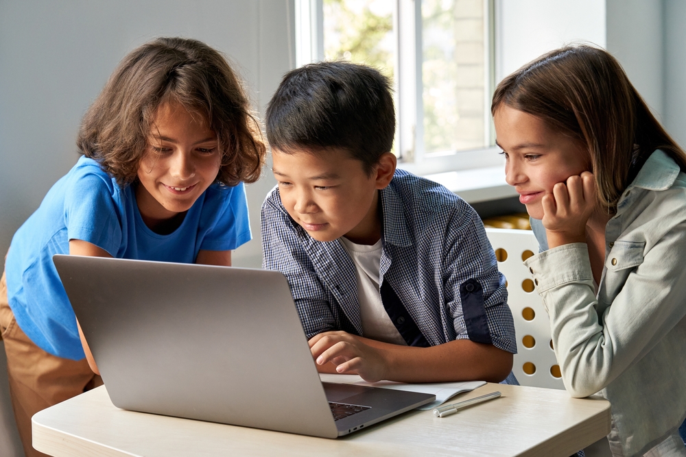Three children are gathered around a laptop on a desk, looking at the screen intently. A pen lies on the desk.